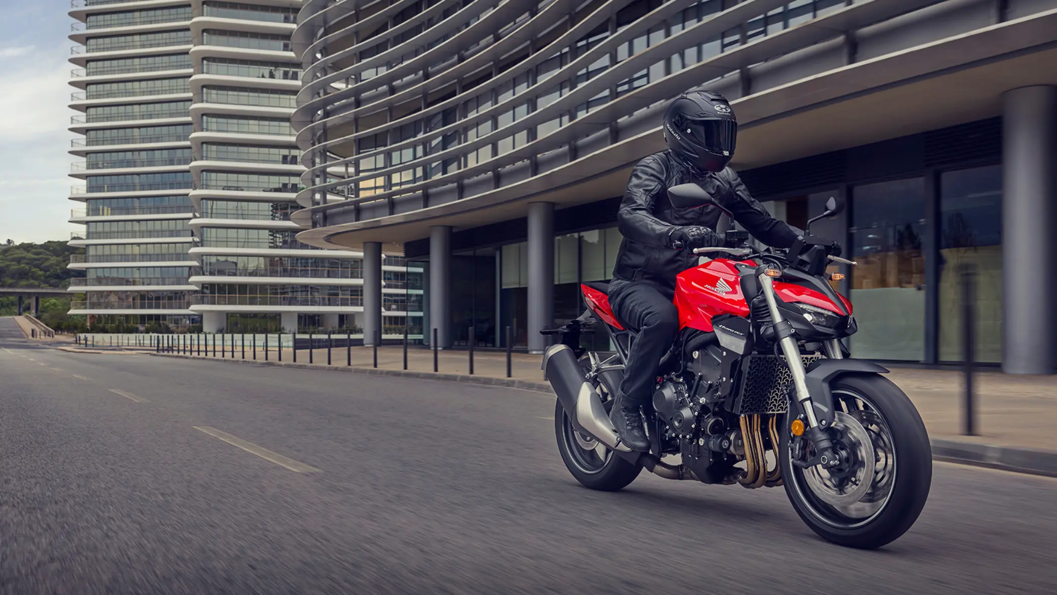 Ein Motorradfahrer auf einer roten Honda CB1000 Hornet fährt auf einer leeren Straße vor modernen Gebäuden.