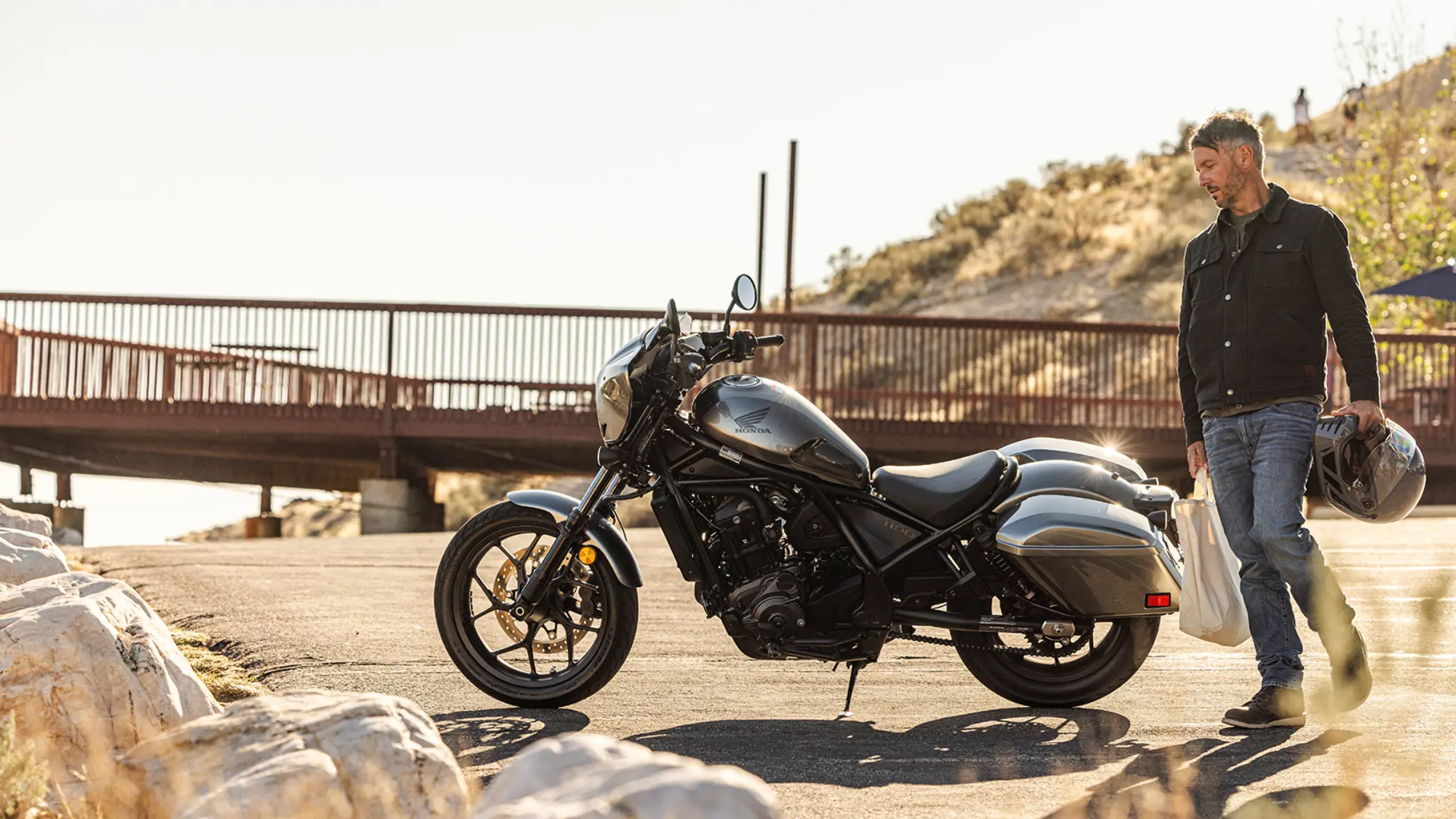 Ein Mann steht neben einem geparkten Honda-Motorrad CMX1100 Rebel und hält einen Helm.