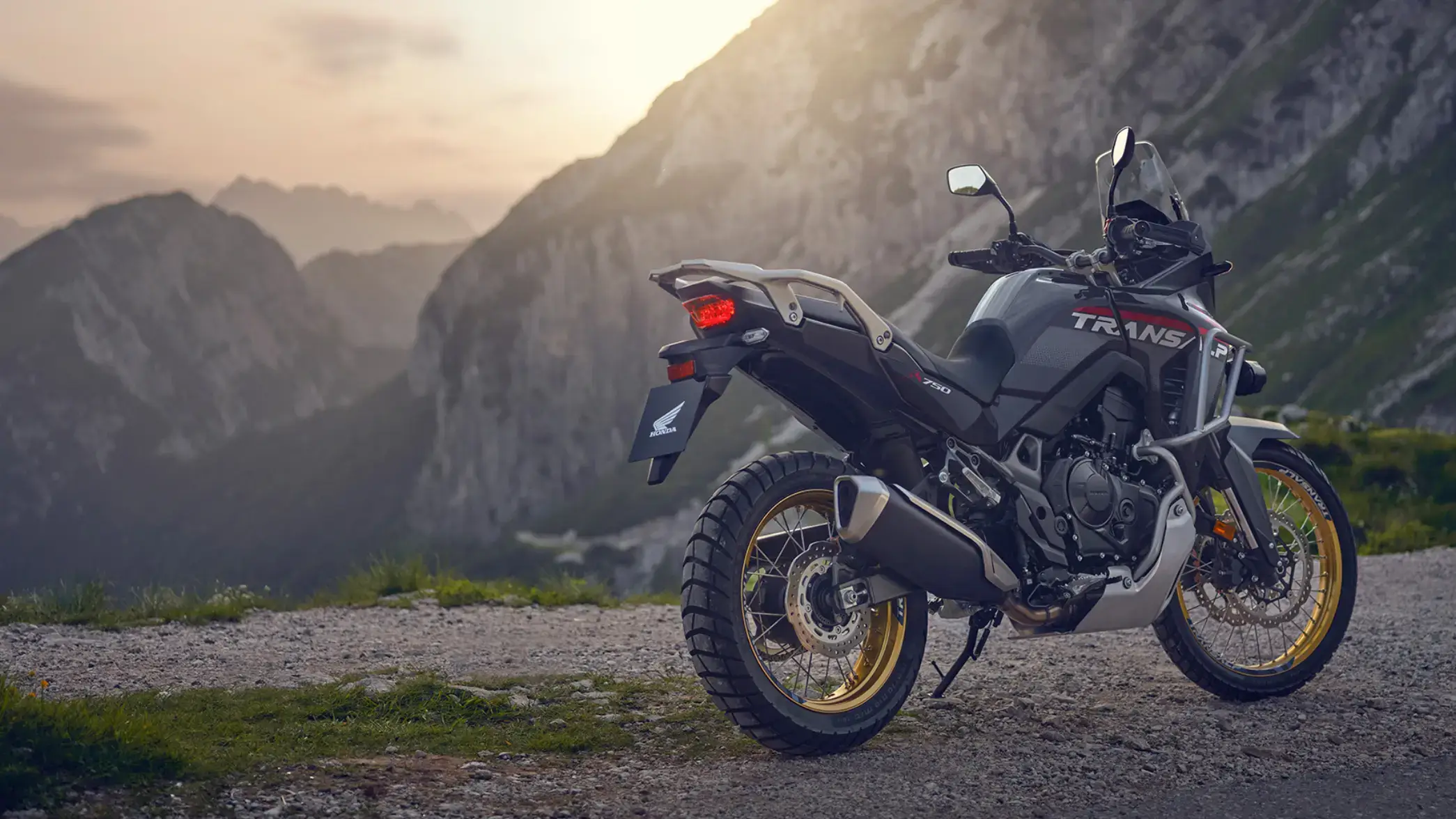 Ein Honda Transalp Motorrad steht auf einem Bergweg mit einer Berglandschaft im Hintergrund.