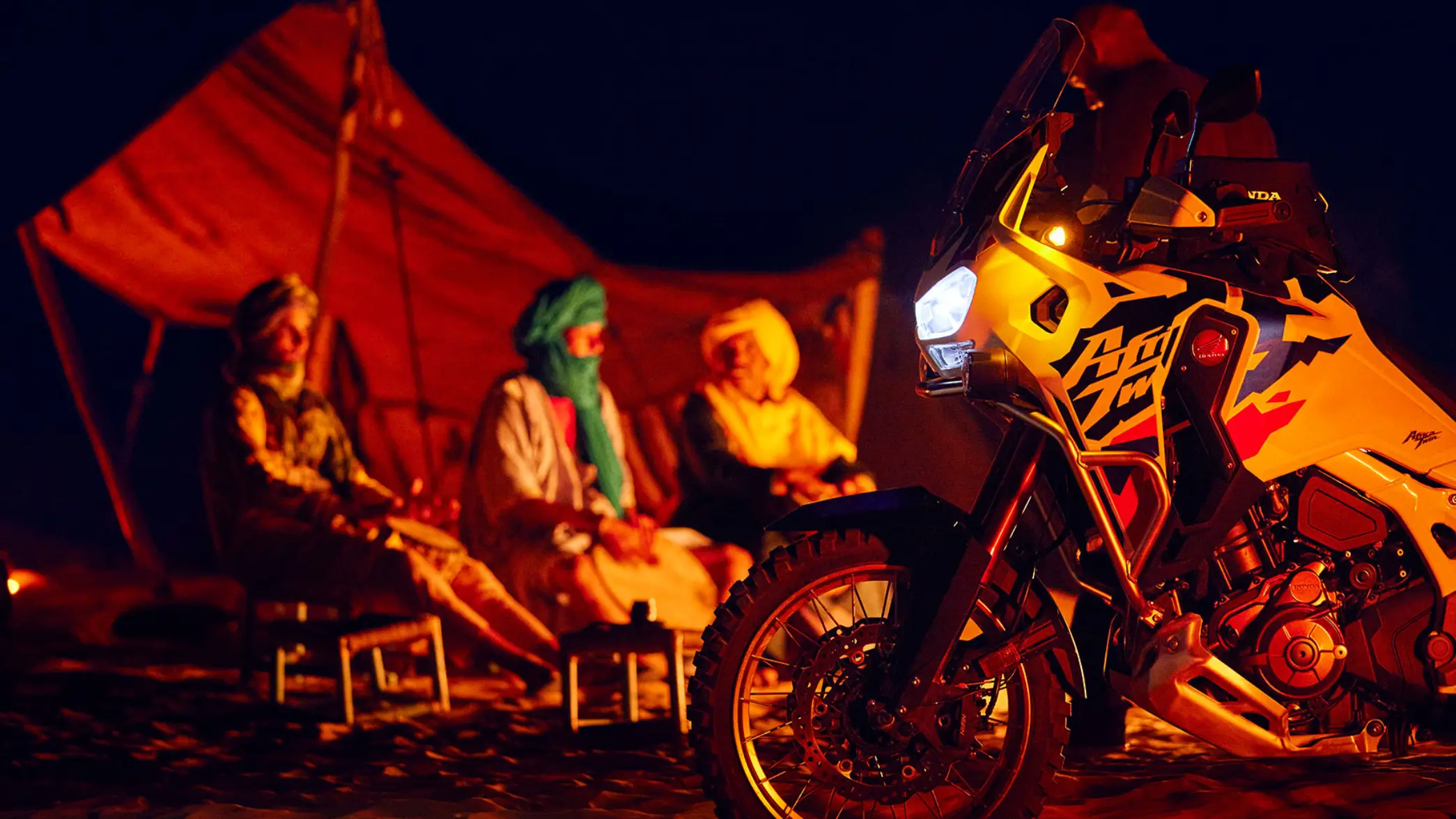 Ein Honda-Motorrad CRF1100L AFRICA TWIN ADVENTURE steht im Vordergrund vor einem Zelt mit drei sitzenden Personen.
