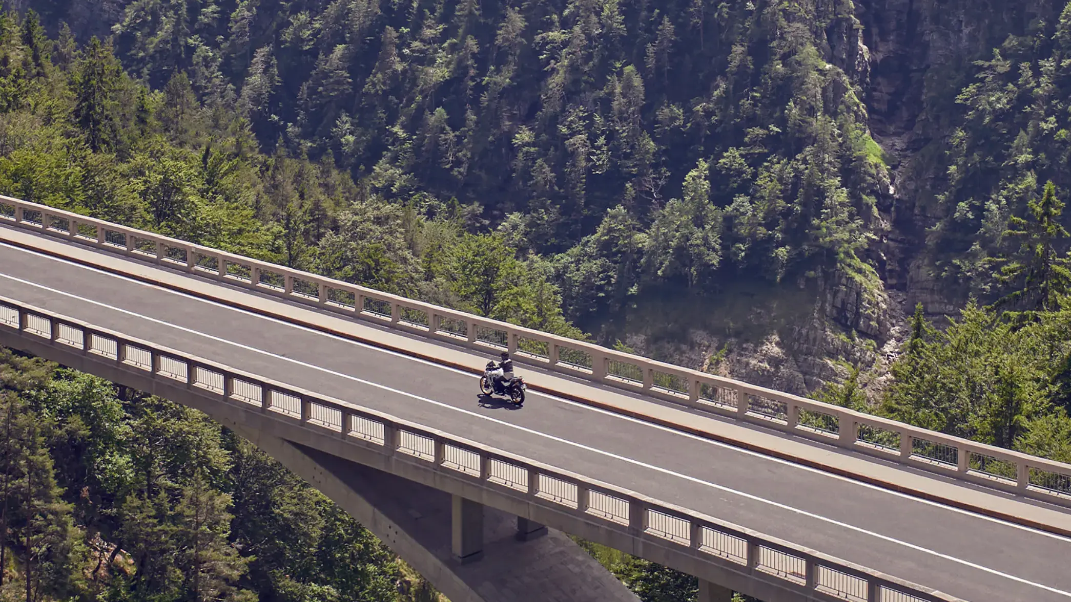 Ein Motorradfahrer fährt über eine Brücke in einer bewaldeten Berglandschaft.