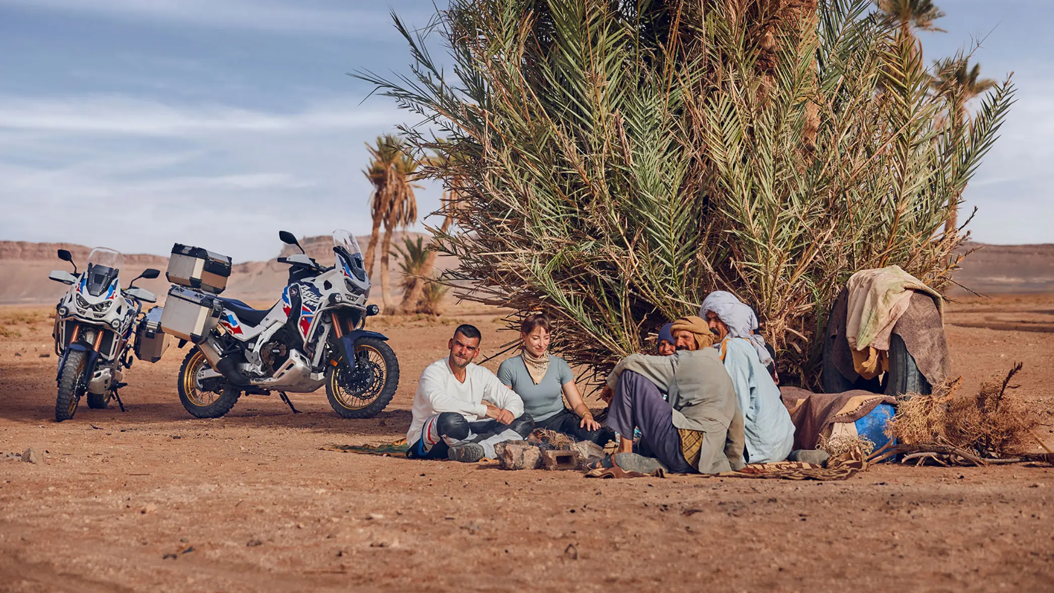 Eine Gruppe von Menschen sitzt neben zwei Honda Africa Twin Motorrädern in einer Wüstenlandschaft.
