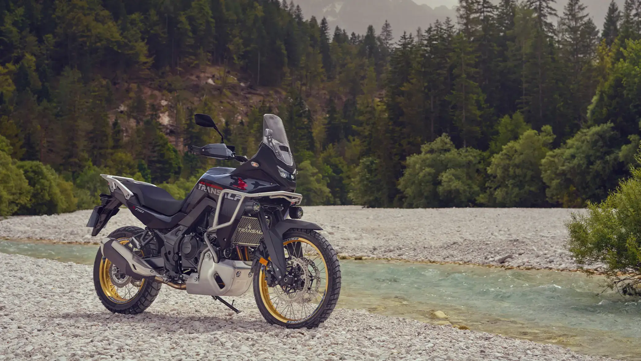 Ein Honda Transalp Motorrad steht auf einem steinigen Flussufer vor einem Wald.