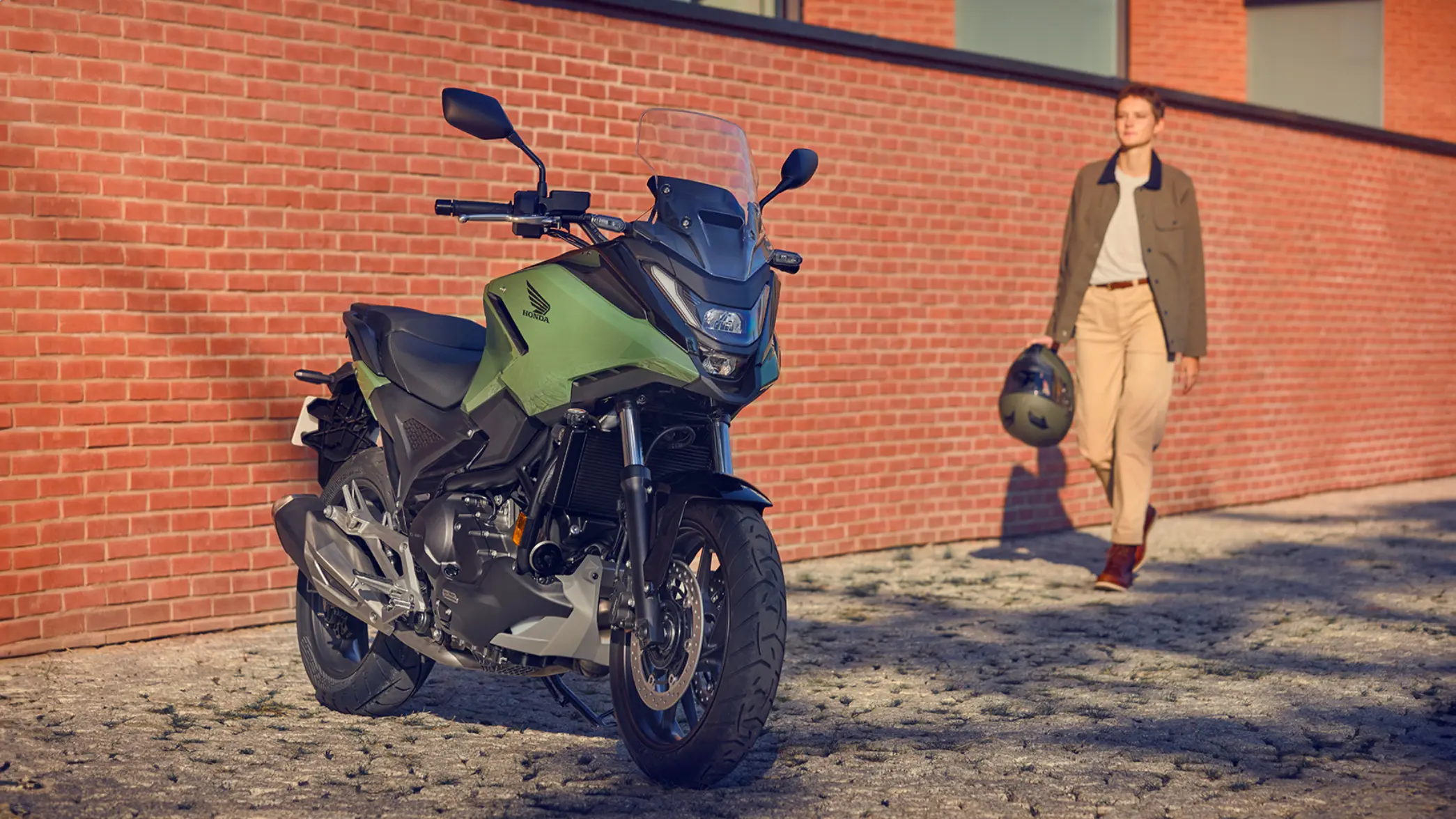 Eine Honda-Motorrad NC750X steht vor einer roten Backsteinmauer, während eine Person mit Helm in der Hand darauf zugeht.
