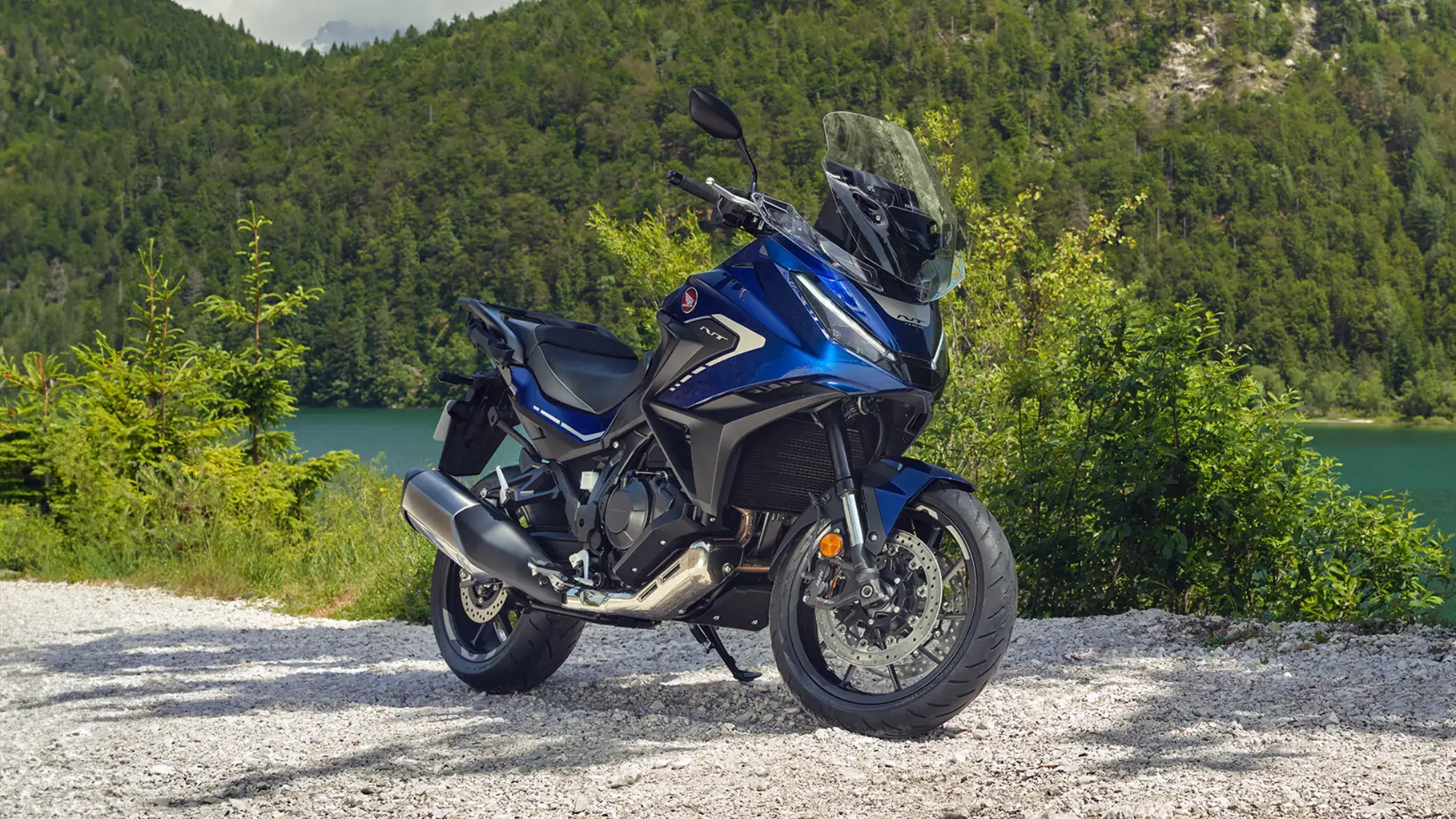 Eine blaue Honda NT1100 steht auf einem Schotterweg vor einer bewaldeten Berglandschaft.