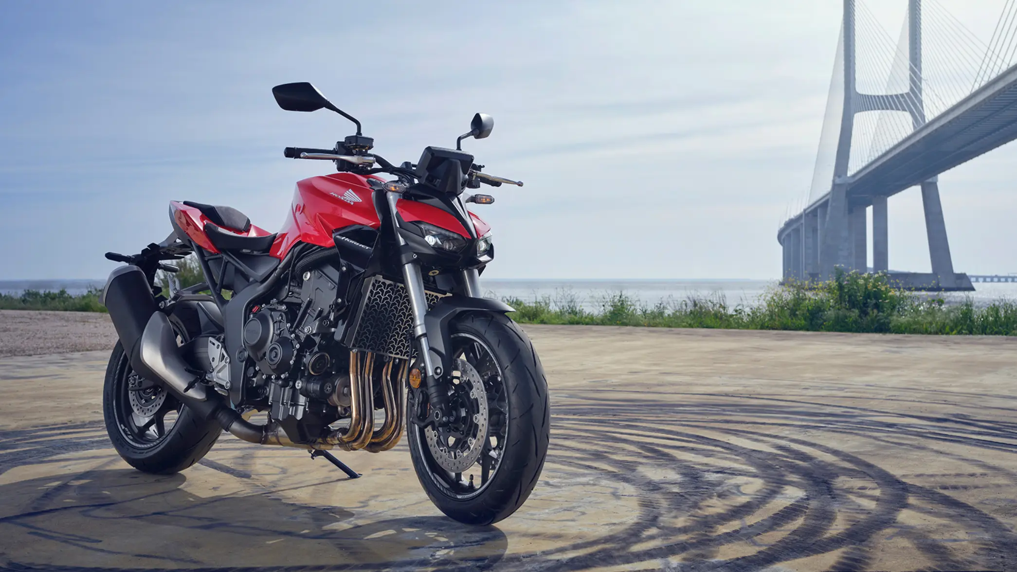 Rotes Honda-Motorrad CB1000 Hornet vor einer Brücke am Meer.