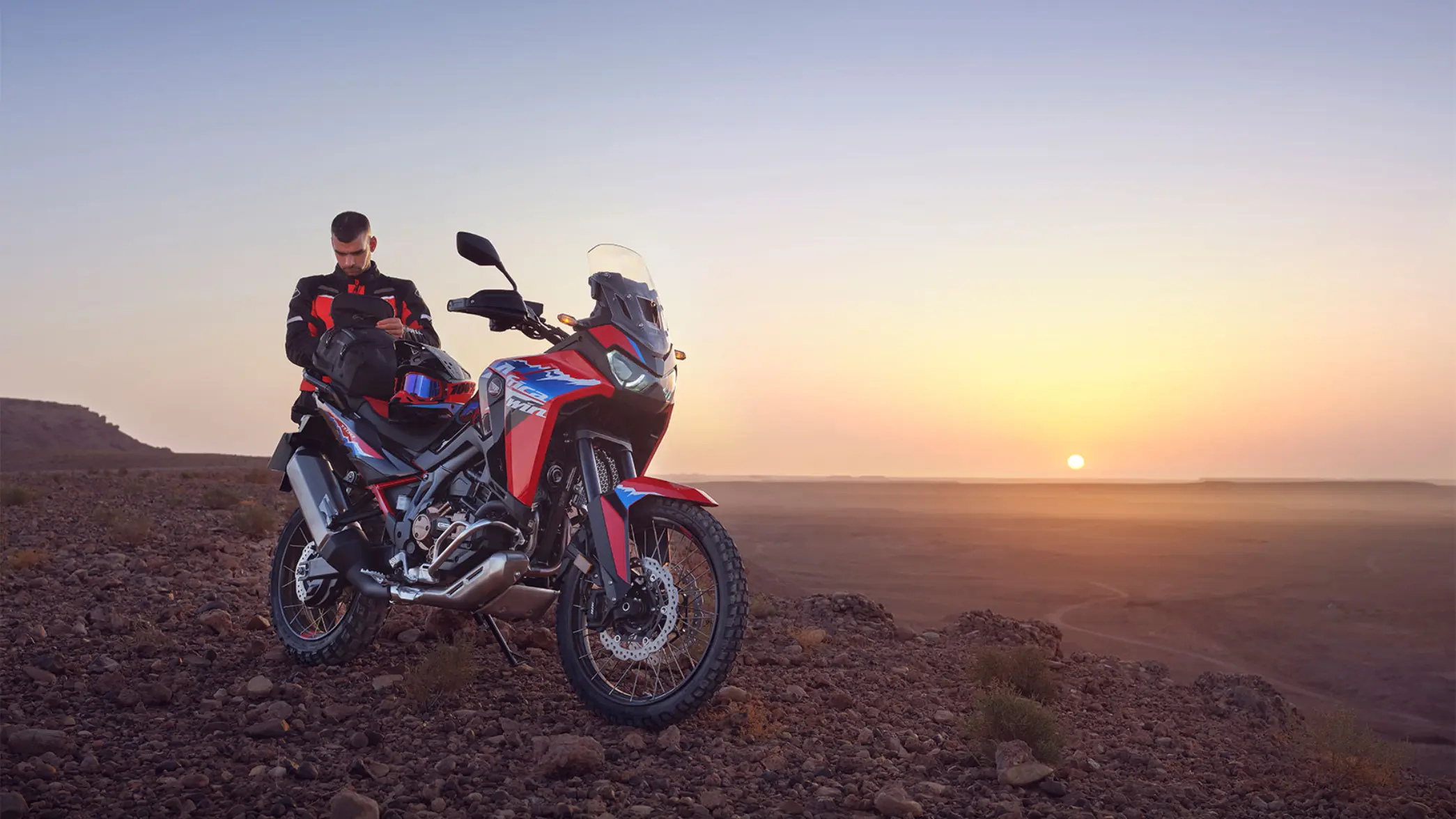 Ein Mann steht neben einem Honda Africa Twin Motorrad in einer Wüstenlandschaft bei Sonnenuntergang.