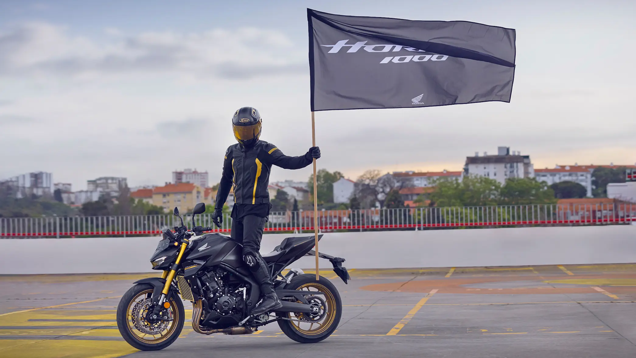 Ein Motorradfahrer steht auf einer Honda Hornet 1000 und hält eine schwarze Flagge mit dem Honda-Logo.
