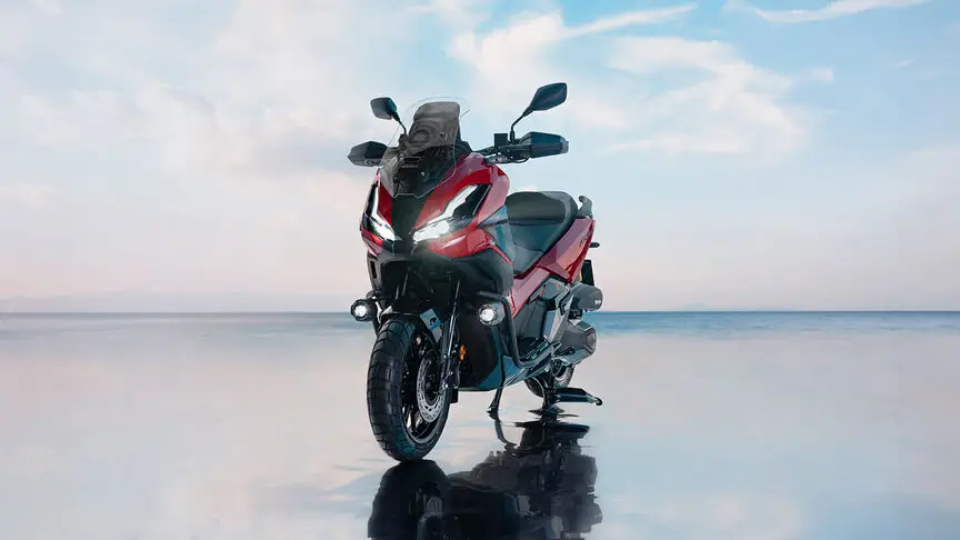 Ein rotes Honda ADV350 Motorrad steht auf einer spiegelnden Oberfläche vor einem blauen Himmel.