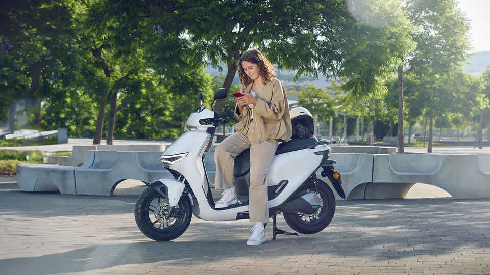 Eine Person sitzt auf einem weißen Honda CUV-e Roller und schaut auf ihr Smartphone.