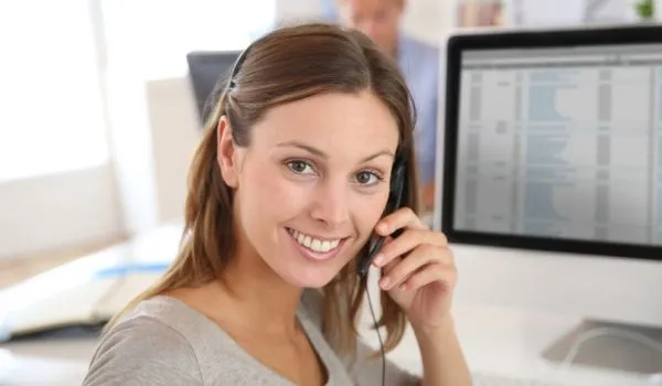 Eine lächelnde Frau telefoniert mit einem Headset vor einem Computerbildschirm.