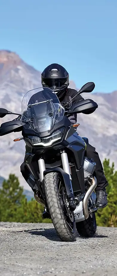 Ein Motorradfahrer fährt auf einer Moto Guzzi Stelvio auf einer Schotterstraße vor Bergkulisse.